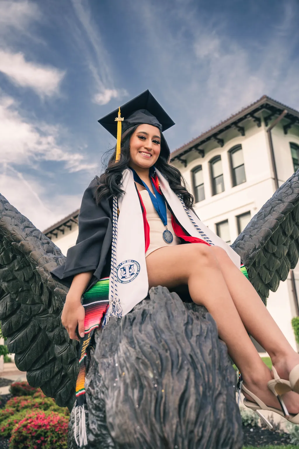 A graduate in cap and gown sitting on the hawk statue at Montclair State University