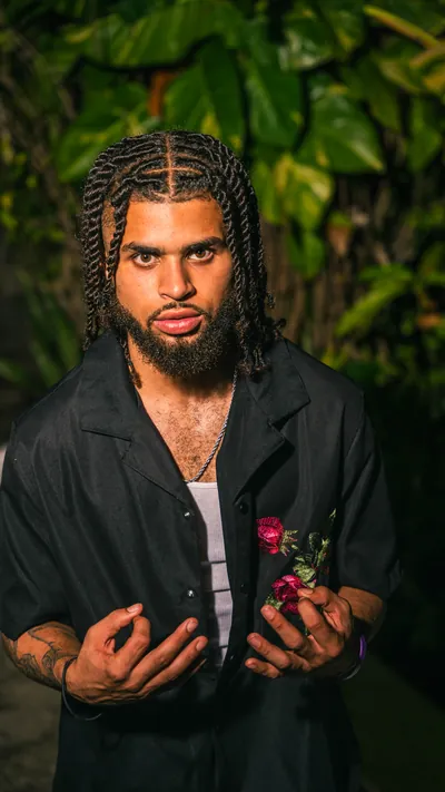Night portrait: a man with braids in a black shirt holding a rose, lit by flash against the leaves