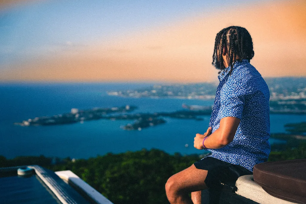 A man in a blue patterned shirt looking out over the sea and the islands at sunset